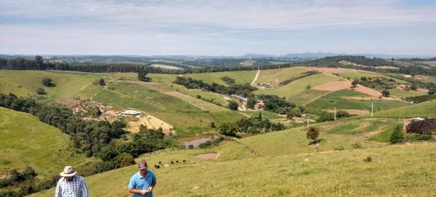 Two people walking uphill in a lush, green rural landscape with rolling hills, scattered farms, and small ponds. The background features distant forests and a partly cloudy blue sky.