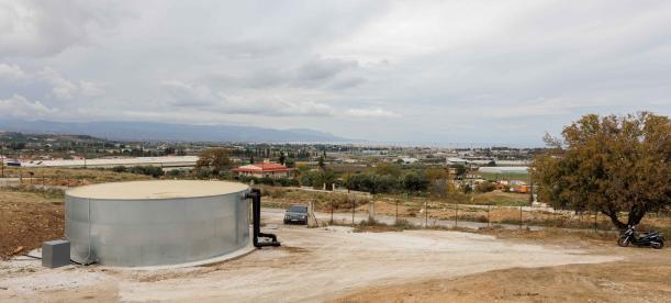 Wide view of a rural landscape with a circular water tank in the foreground, surrounded by dirt paths and sparse vegetation.