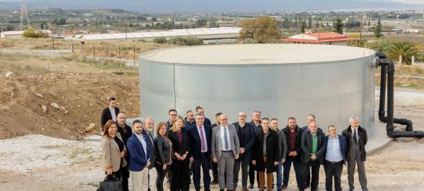 A group of people posing in front of a circular water tank in an outdoor setting, with a rural landscape and distant mountains in the background. The individuals are dressed in professional attire.